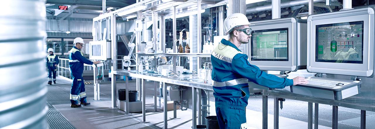 [photo] Man wearing glasses, hardhat, and heavy clothing, standing in front of two large computer screens in a manufacturing facility
